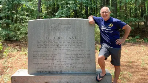 Sean Drysdale Sean Drysdale who is bald on top and has grey hair at the side and is wearing a Norn Iron blue T-shirt and grey shorts stands beside a a stone sign that says Old Belfast with a forest in the background