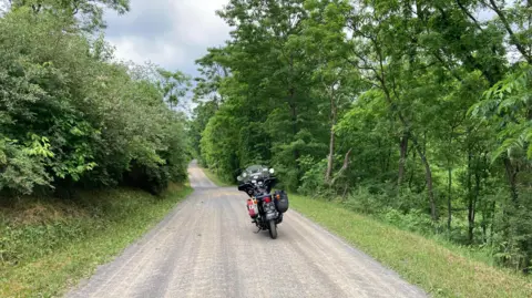 Sean Drysdale A motorbike parked in the middle of a forest road with green trees and bushes overhanging the narrow road