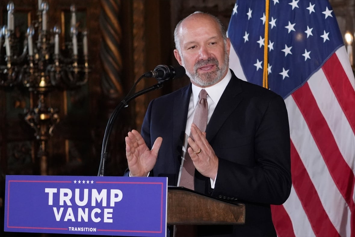 Howard Lutnick speaks at a podium labelled 'Trump Vance Transition,' with the U.S. flag in the background.  