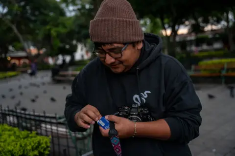 Fritz Pinnow Ronald Ottoniel, wearing a woolly hat and thick-rimmed glasses, is loading his new film roll into his camera ay a square in Antigua. Pidgeons can be seen on the ground in the background.