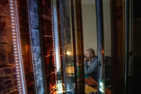 Fritz Pinnow Fabriccio Díaz is working on his computer in room of their apartment that has been converted into an office. In the foreground, rolls of film are hanging to dry. 