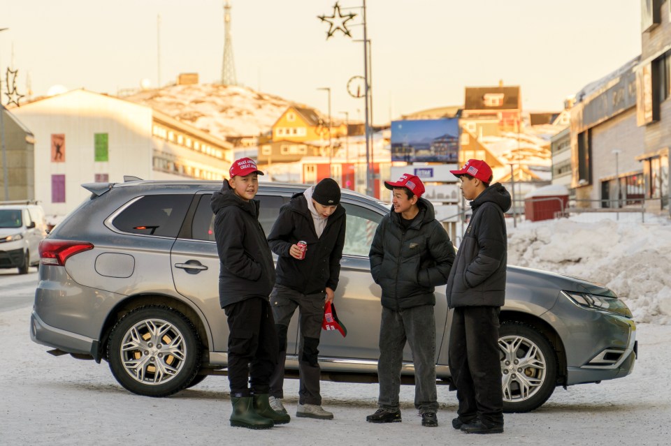 Four boys wearing red hats stand by a car in a snowy town.