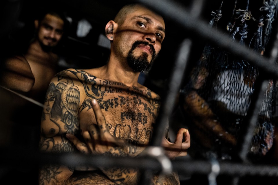 A heavily tattooed MS-13 gang member in a crowded jail cell makes a gang sign.