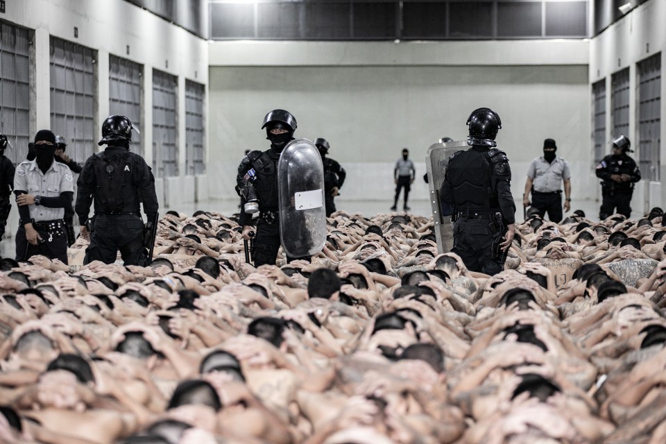 Gang members with their hands on their heads being transferred in a prison, guarded by police.