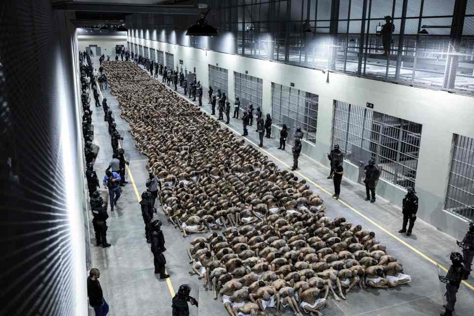 Hundreds of shirtless gang members sit on the floor of a prison, surrounded by guards.