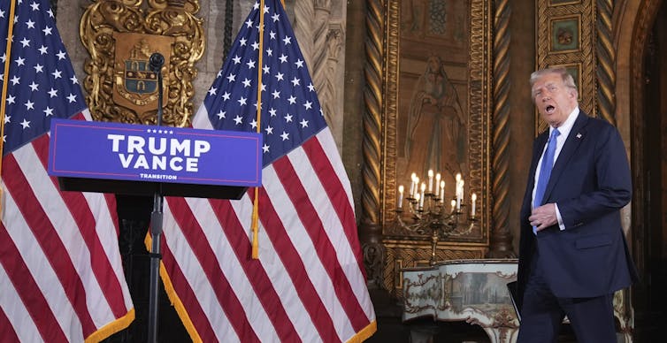 An older rotund man with his mouth open and fluffy grey-blond hair walks towards a lectern.