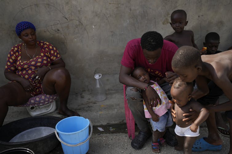 A Black family with several small children sit in the shade outside a concrete structure.