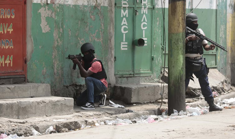 Two heavily armed police officers, one crouching and the other standing behind a pole. Both wear helmets.