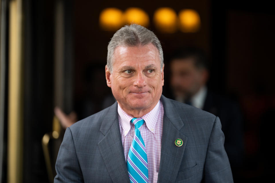 A man in a suit with a striped tie stands outside a building, looking to the side with a slight smile