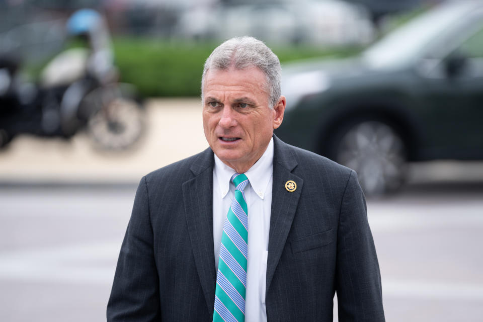 Carter in a suit and striped tie stands outside with cars and a motorcycle in the background