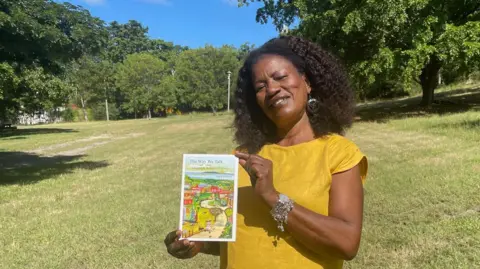 Gemma Handy Joy Lawrence, surrounded by grassland and trees, holds up her book The Way We Talk