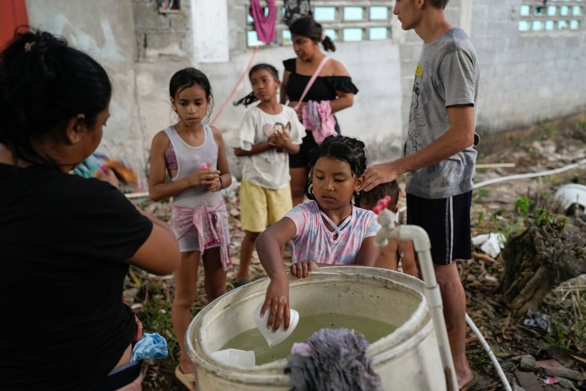  A child puts their hand with a plastic cup into a large pail, with a few children lined up behind.