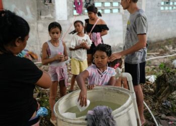 A child puts their hand with a plastic cup into a large pail, with a few children lined up behind.