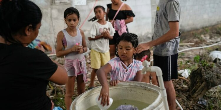 A child puts their hand with a plastic cup into a large pail, with a few children lined up behind.