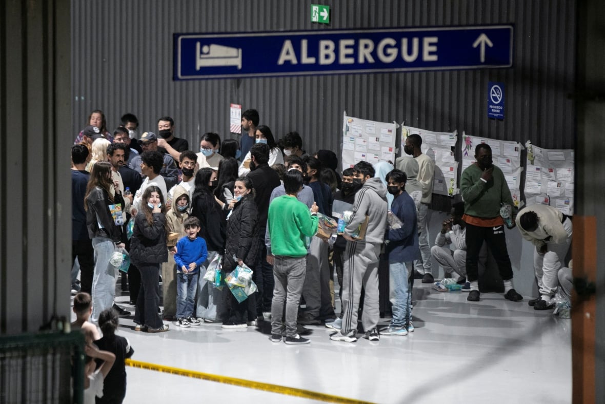 A tightly packed-in crowd of dozens of people are shown inside an airport terminal from an elevated view.