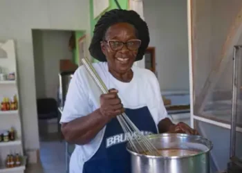 Gemma Handy Novella Payne, wearing glasses and a hair covering, stirs a pot containing a sauce she makes using traditional recipes