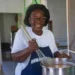 Gemma Handy Novella Payne, wearing glasses and a hair covering, stirs a pot containing a sauce she makes using traditional recipes