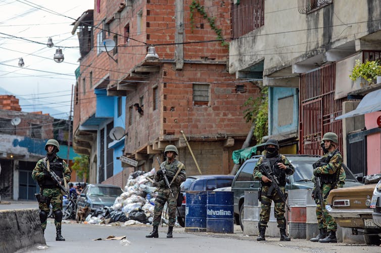 People in army fatigues and holding guns stand in front of a building.