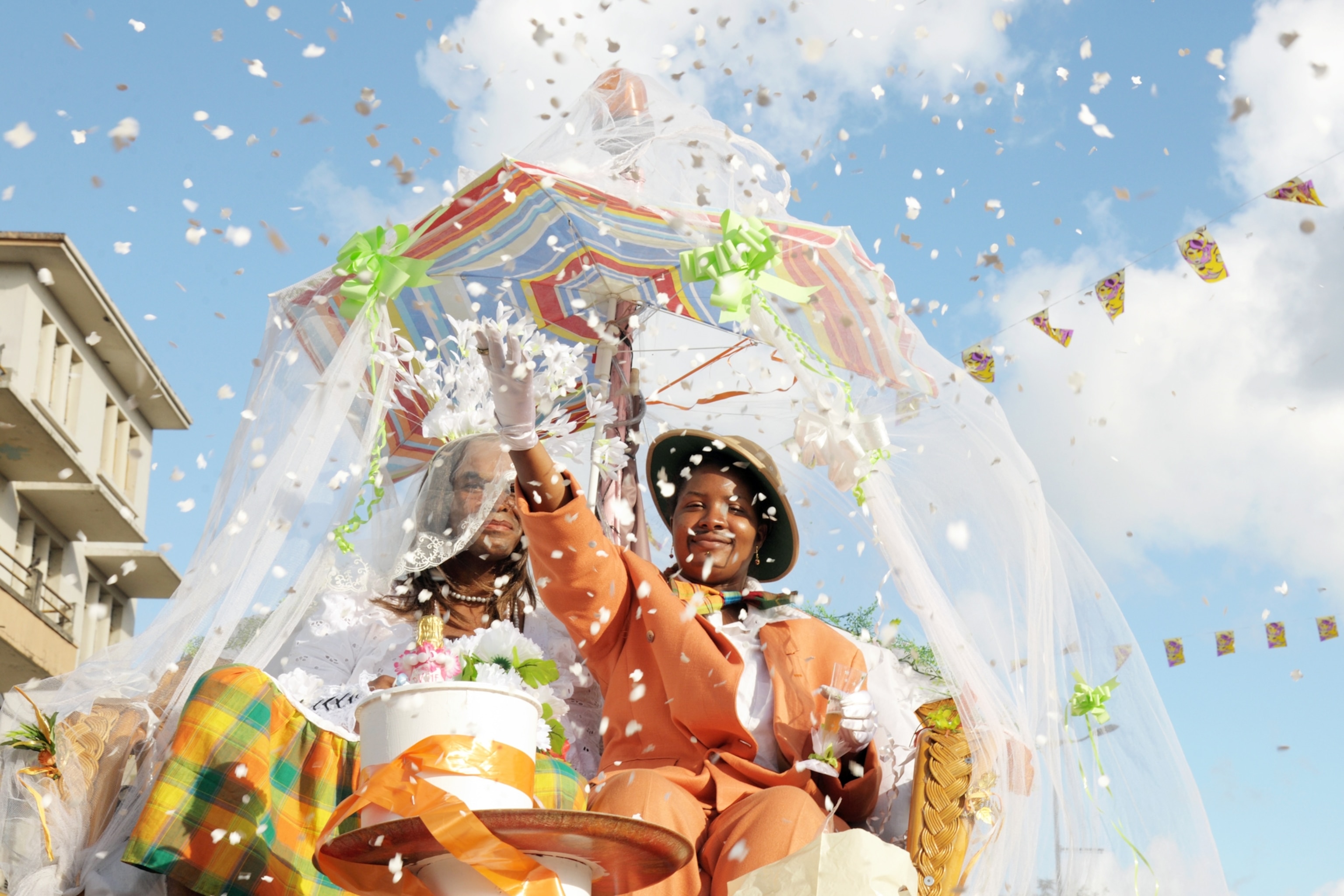 Confetti falls from the sky as people celebrate in costume with parasols under a blue sky.